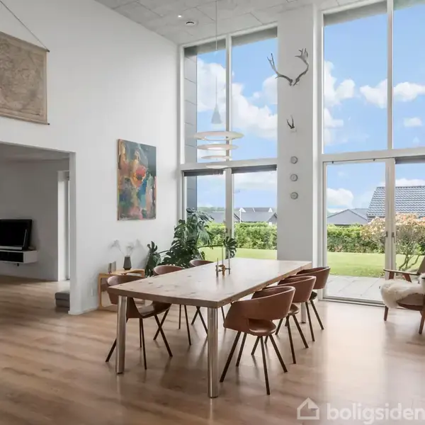 Et stort spisebord med seks stole står i et lyst rum med gulv-til-loft-vinduer. Udenfor ses en have. En stue med sofaer og fjernsyn er i baggrunden.