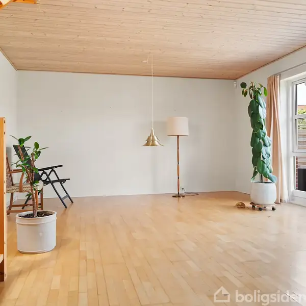 Et lyst rum med trægulv og trappe, indeholder to potteplanter og en hvid lampe. Store vinduer viser en terrasse udenfor. To stole står i hjørnet.