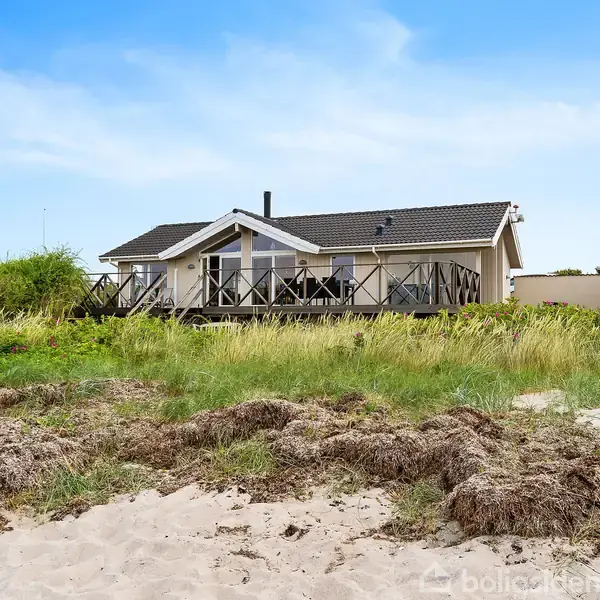 Et sommerhus med terrasse ligger stille ved en strand. Sand og græs dækker jorden omkring huset, som har en moderne design i en naturskøn kystomgivelser.