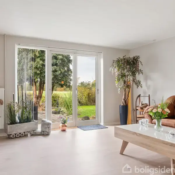 En stue med en brun lædersofa står op ad en væg ved siden af et sofabord med blomster. Store vinduer viser en have med grønt græs og buske udenfor.