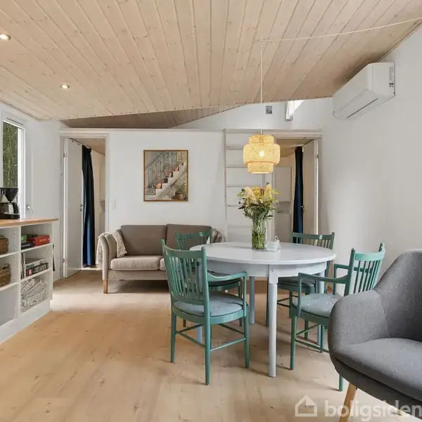 En stue med et rundt spisebord omgivet af fire stole. Blomster står som borddekoration. Sofa i baggrunden, omgivet af lys træindretning og grøn udsigt gennem vinduerne.