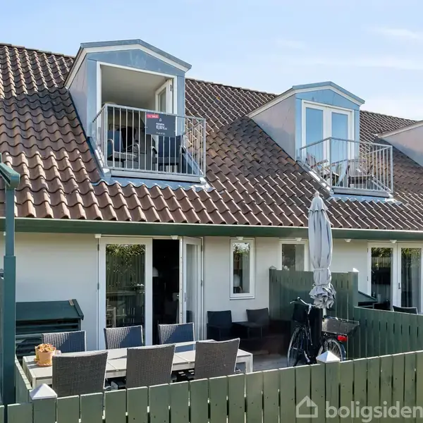 Rækkehus med rødt tegltag og flere kviste, stille i en række. Foran er en terrasse med havemøbler og parasol, omgivet af grønne hegn, i solrigt boligområde.
