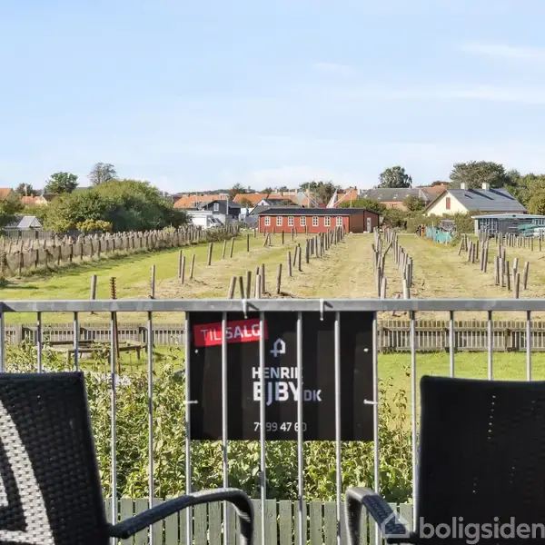 To stole står på en altan med udsigt over en vingård, der slynger sig gennem et landligt område. Skilt med teksten "Til salg" og "Henrik Br. B. 7639 4747".