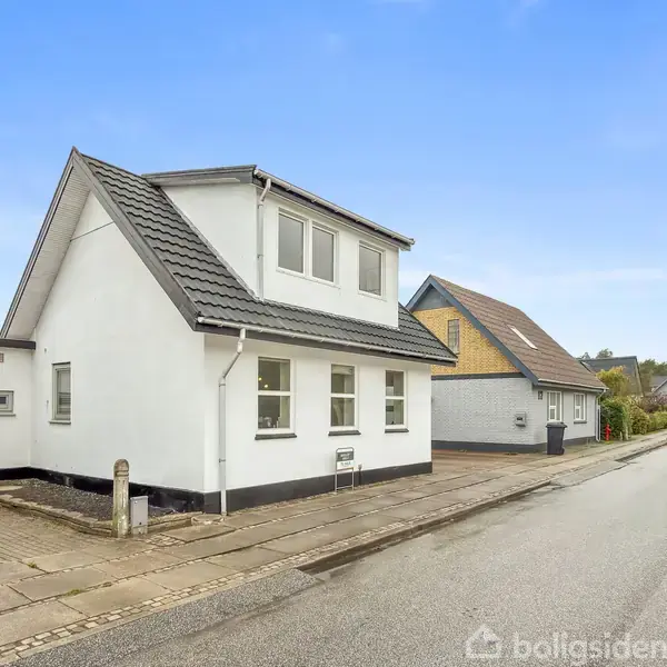 Hvidt hus med gråt tag står stille ved en rolig vej i et boligområde. To etager med flere vinduer. Andrids kompasretning indikerer vest.