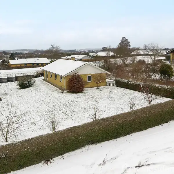 En gul bygning står i en snedækket have omgivet af hække. Buske og et skur ses i baggrunden, med flere huse synlige i horisonten under en klar, blå himmel.