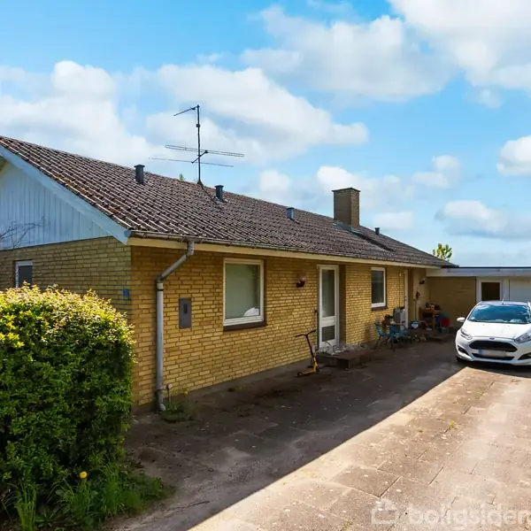 Huset er en gulstensbungalow med skråt tag, placeret ved en flisebelagt indkørsel, hvor en hvid bil holder parkeret. Omgivet af grønne buske og under en blå, skyet himmel.