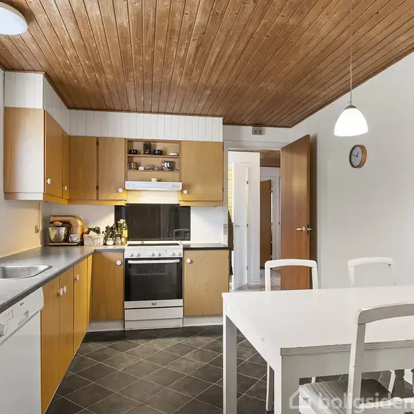 Et køkken i træ og hvidt med et komfur og ovn, en hvid vask, en opvaskemaskine til venstre, og et hvidt bord med to stole i forgrunden. En dør og ur er på væggen.