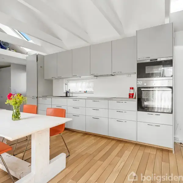 Køkkenet har en hvid bordplade og grå kabinetter. På bordet står en vase med blomster. Fire orange stole omgiver bordet. Trægulv og loft med ovenlys skaber en lys atmosfære.