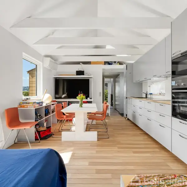 Stue med moderne køkkenindretning, hvor et spisebord med orange stole er central. Hylder, og TV placeret i en lys rum med trægulve. Store vinduer giver udsigt til haven.