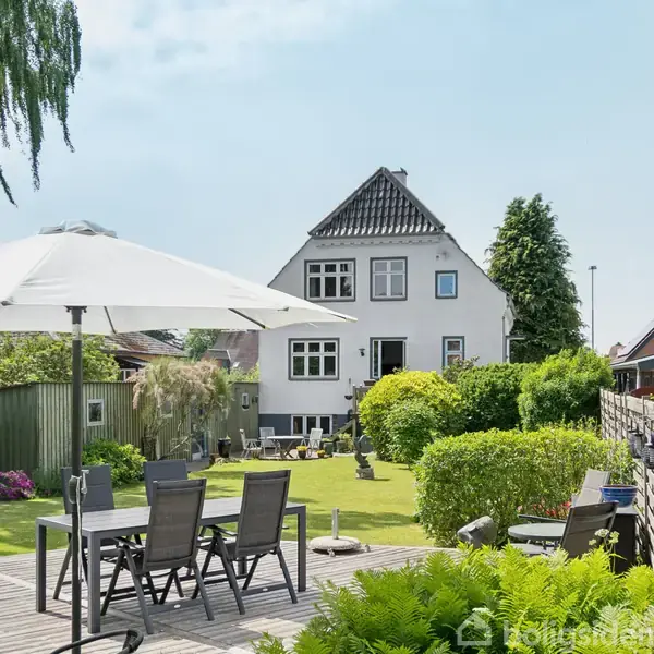 Et hvidt hus med spidst tag står i baggrunden. På en terrasse foran er der havemøbler og en stor parasol. Haven er frodig med buske og træer omkring.