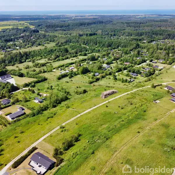 Grønt landskab set fra oven med spredte huse, der hviler i rolige landlige omgivelser. Veje snor sig gennem området, omgivet af træer og marker under en blå himmel.