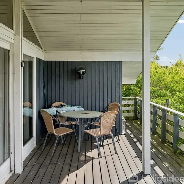 En overdækket terrasse med to kurvestole og et bord står op ad en mørk trævæg. Til højre er grønne træer og en udsigt over landskabet.