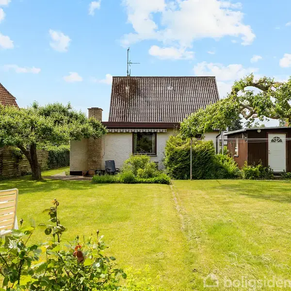 Et lille hus står stille i en grøn have med frodige buske og træer. En hvid bord-bænk-sæt er placeret på græsplænen, omgivet af solskin.