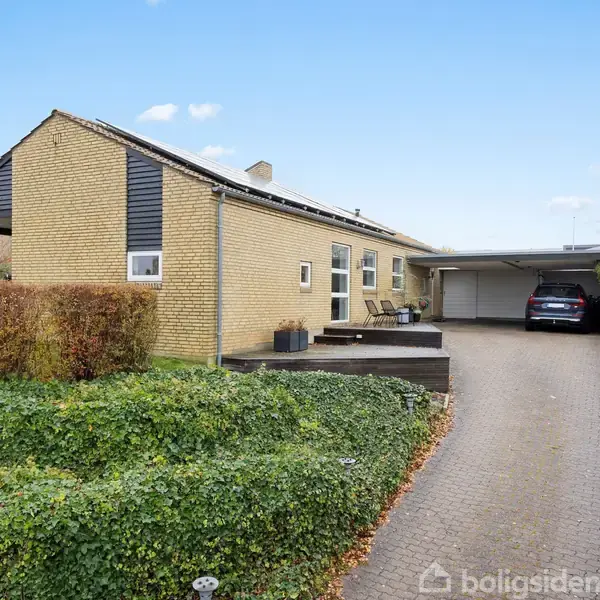 Et gult murstenshus med flere vinduer står for enden af en indkørsel. En bil holder parkeret under en carport. Omgivet af grønne buske og hække.