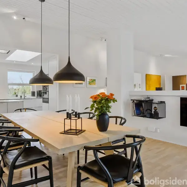 Et træbord omgivet af sorte stole står centralt i et moderne, lyst køkken med hvide vægge. Over bordet hænger sorte pendellamper. Blomster i en vase pryder bordet.