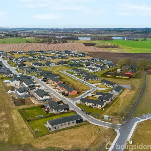 Boligområde med moderne huse ligger i et åbent landskab, omgivet af marker og grønne områder. Veje forbinder husene, og terrænet er fladt med spredte træer omkring.