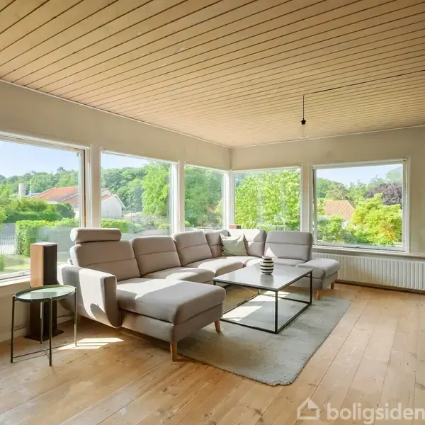 En stor, lys stue med en lysegrå hjørnesofa står på et trægulv. Sofaen er placeret ved store vinduer, der viser en grøn have udenfor. Fjernsynet hænger på væggen modsat sofaen.