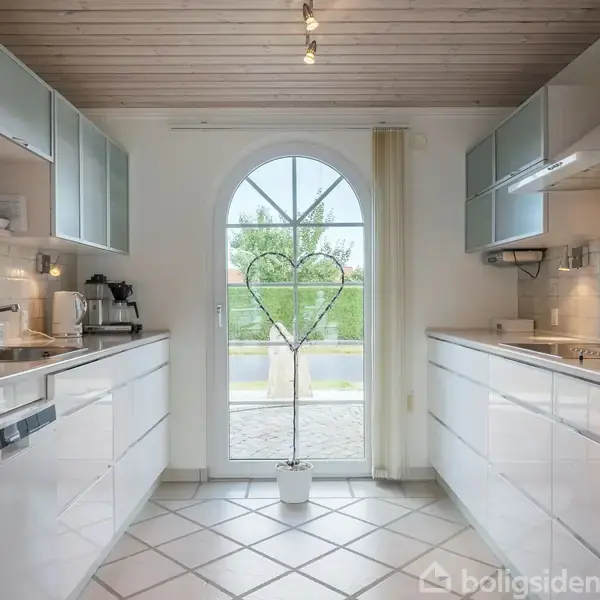 Et lyst køkken med hvide skabe og apparater arrangeret langs begge sider. Et stort, buet dørparti med glas åbner ud til en terrasse. En potteplante står foran døren.