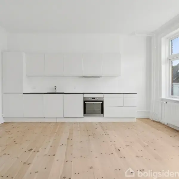 Hvidt køkken står ubenyttet med indbygningsovn og vask i et lyst rum med trægulv, store vinduer til højre giver naturligt lys.