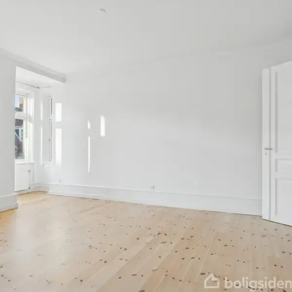 Et tomt, lyst rum med trægulv, hvidmalede vægge og store vinduer, der slipper sollys ind. En åben hvid dør fører til et andet rum.