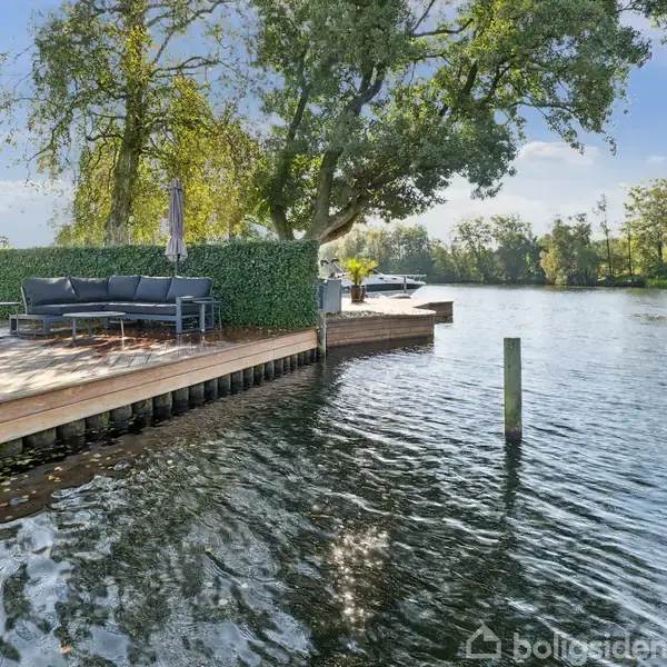 En terrasse med udendørsmøbler står ved en søbred. Træer omkranser området, og himlen er blå. Møblerne inkluderer en stor sofa og et bord.