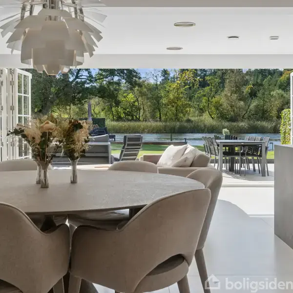 Spisebord med blomster står i en lys stue med åbne døre til en terrasse. Udenfor ses en grønt område med træer. Der er spisebordsstole udenfor.