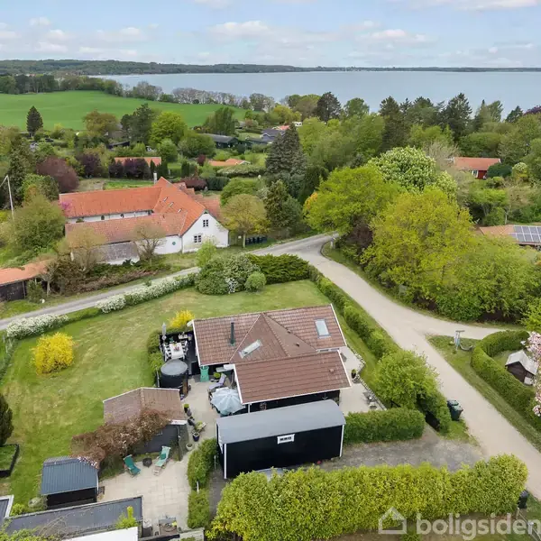 Hus set ovenfra med rødt tag, beliggende i en frodig have med træer og buske, omgivet af en vej. I baggrunden ses landskab med grønne marker og en sø.