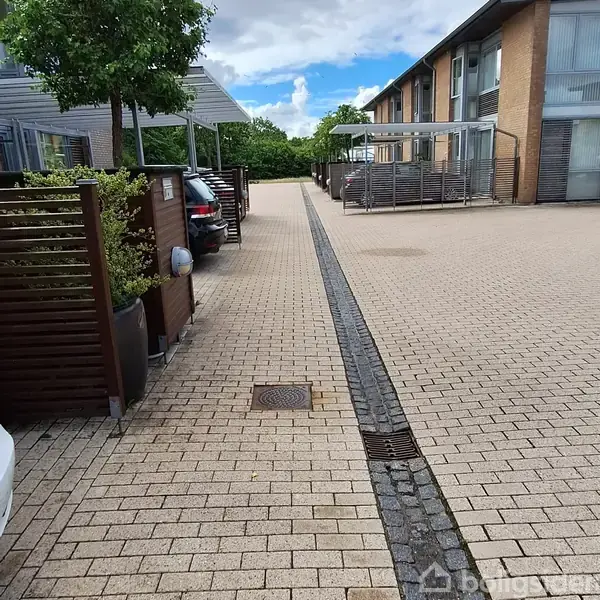Parkerede biler står under halvtag langs en bred flisebelagt vej. Vejen har en afvandingsrende i midten, og omgivelserne inkluderer moderne bygninger og grønne træer i horisonten.