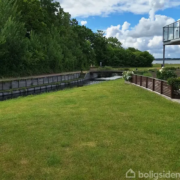 En grøn græsplæne strækker sig langs en stille kanal med træpaneler. Træer og buske udgør baggrund, mens en bygning med balkon er synlig til højre.
