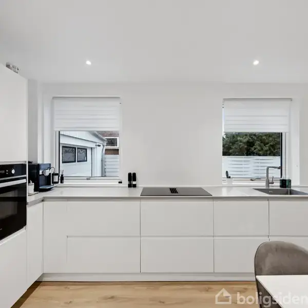 Køkken med hvide skabe har en indbygget ovn og kogeplade. To vinduer lader naturligt lys komme ind. Grå stole ses i forgrunden, trægulv skaber en varm atmosfære.