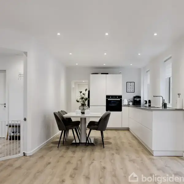 Spisebord med fire stole står i et moderne, lyst køkken med hvidt interiør og træparketgulv. Der er indbyggede skabsapparater og en stor køkkenø med blomster i en vase.