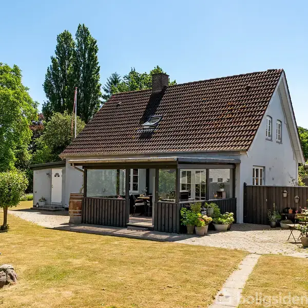 Hus med rødt tegltag står i en solrig have. En træveranda er foran huset, omgivet af græsplæne, blomsterkrukker og stensti. Store, grønne træer i baggrunden.