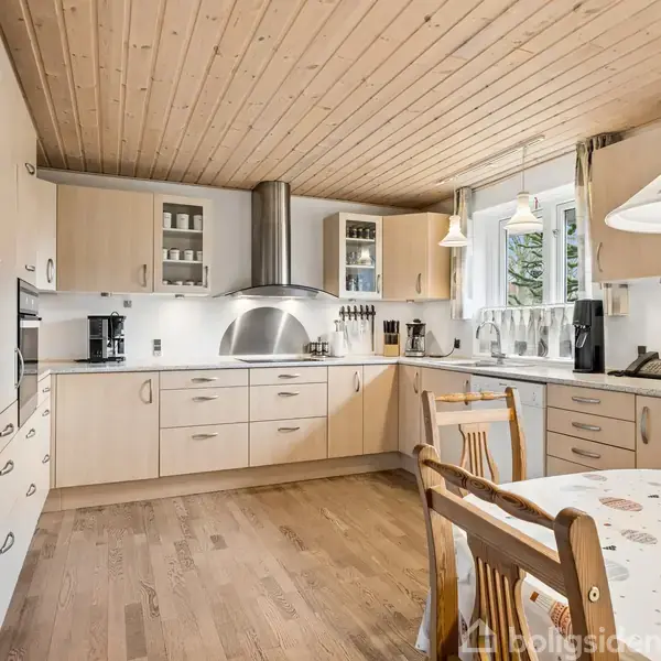 Køkken med træloft og træmøbler, herunder et spisebord med dug, moderne køkkenudstyr og apparater, bl.a. ovn og opvaskemaskine, vinduer med udsigt til grønne omgivelser.
