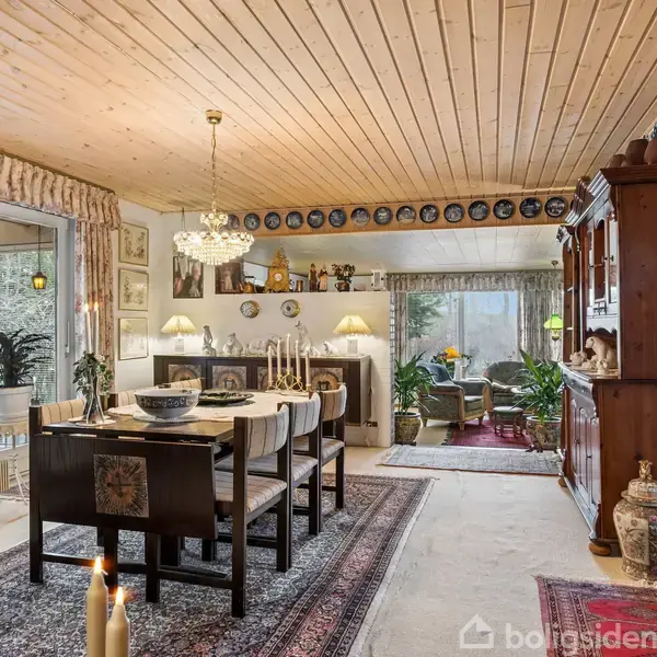 Spisestue med træloft og stor lysekrone. Bordet er dækket med stole omkring. Omgivet af billeder på væggene, planter og porcelænsdekorationer, med tæppe på gulvet.