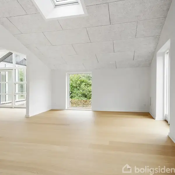 Et lyst, tomt rum med træparketgulv og hvide vægge. Loftet har et ovenlysvindue. Store vinduer giver udsigt til en grøn have. Rummet leder til en anden del af huset.