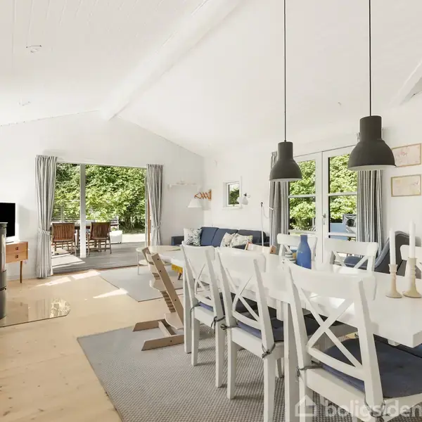 Spisebord med hvide stole står i en lys stue med trægulv. En sort brændeovn er tændt. Store vinduer og en åben dør viser en terrasse med grønne omgivelser udenfor.