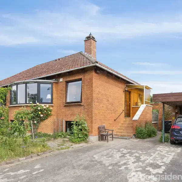 Rødt murstenshus med højt tag og skorsten, omgivet af have. En bil står parkeret under en carport mærket med nummeret 10. Trappe fører til en hoveddør.