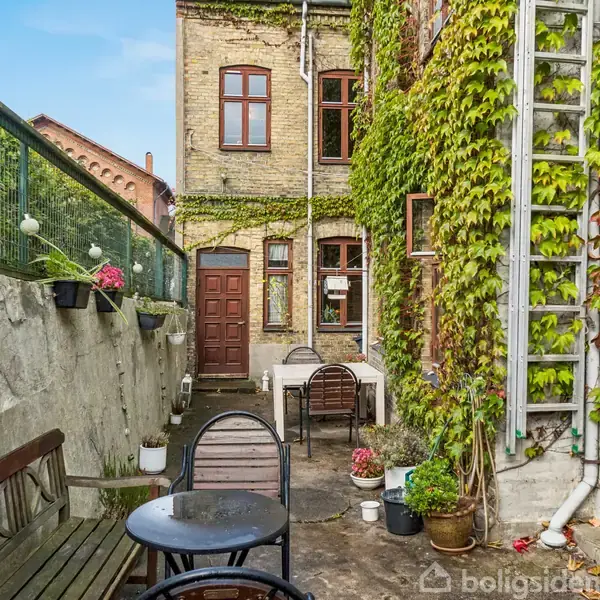 Bord og stole står i en hyggelig baggård med en murstensbygning dækket af grønne planter. En havelåge er åbnet, blomster hænger i potter, og en stige er op ad muren.