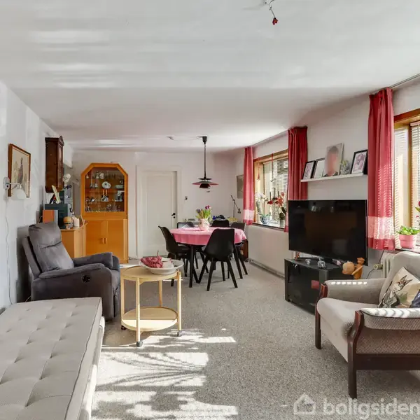 Stue indrettet med sofaer, lænestol og kaffebord, flankeret af en spiseplads med rødt bordklæde. Rummets vægge er dekoreret med malerier; vinduer med røde gardiner giver naturligt lys.