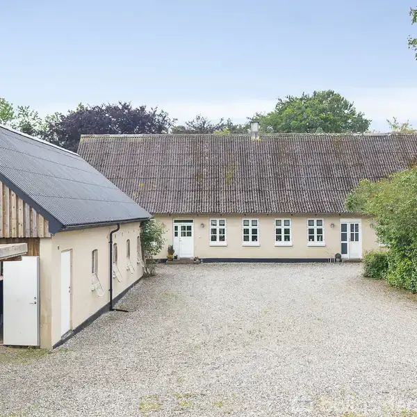 Et traditionelt hus står foran en grusbelagt gårdsplads omkranset af et grønt område; en bil holder til venstre ved en bygning med træbeklædt facade.