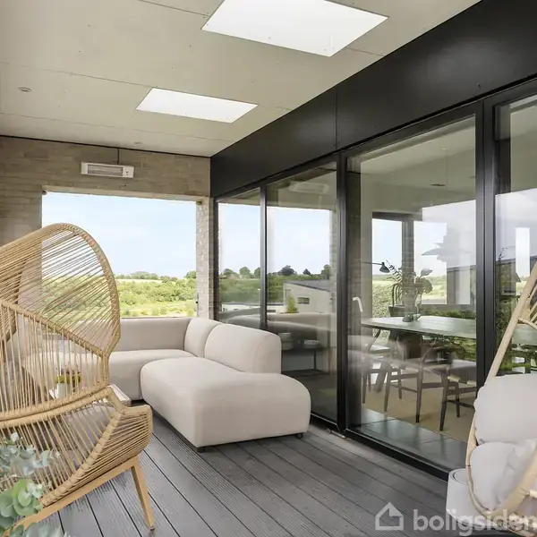 Terrasseområde med to hængestole i bambus hængende fra loftet. Beige sofa står ved ruderne, der adskiller rummet fra den grønne udsigt udenfor. Trægulv og indrammede glasfacader.