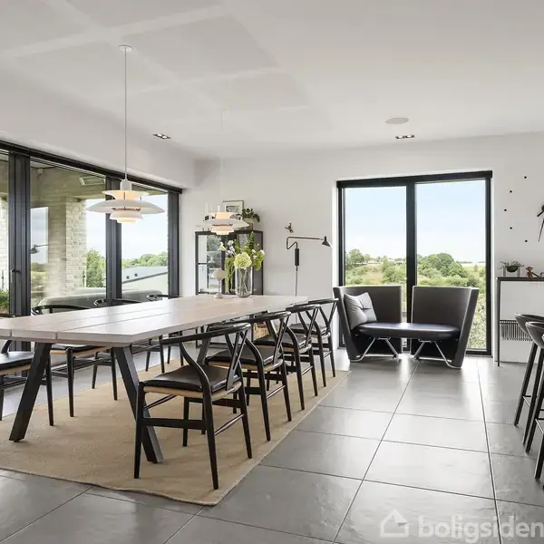 Et spisebord med sorte stole står i et moderne, lyst køkken-alrum. Store vinduer åbner mod en terrasse med udsigt over grønne omgivelser. En veldesignet lampe hænger over bordet.