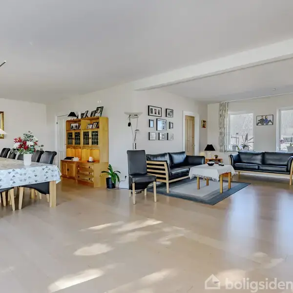 Stue med spisestue og opholdsområde. Et spisebord med blådug og stolopstilling står til venstre, mens sorte sofaer og et sofabord på tæppe ses til højre. Rummelige vinduer bagtil.