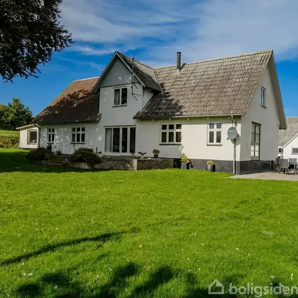 Hus omgivet af grøn græsplæne og træer, står stille under en blå himmel. Der er en terrasse med stole tæt på bygningen.