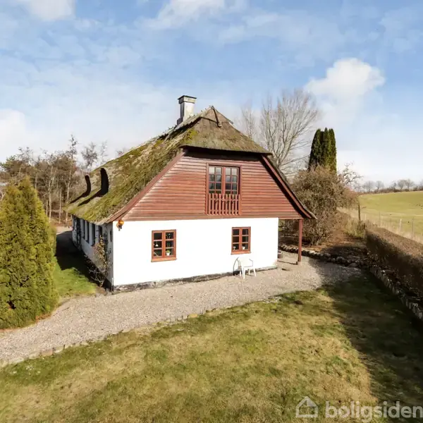 Hus med stråtag står stille omgivet af grøn græsplæne og buske, ved siden af en grusvej. Huset har hvide vægge, træpaneler på gavlen og en klar blå himmel bagved.