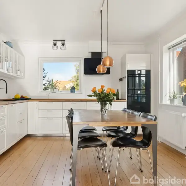 Et moderne køkken med hvide skabe og sort vask. Et bord med sorte stole står centralt, dekoreret med en buket orange blomster. Store vinduer slipper lys ind.