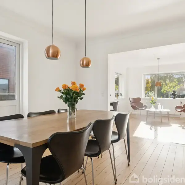 Et træbord med seks sorte stole står i et moderne, lyst spisestueområde. En vase med orange roser står på bordet under to kobberfarvede lamper. Stuen ses i baggrunden.