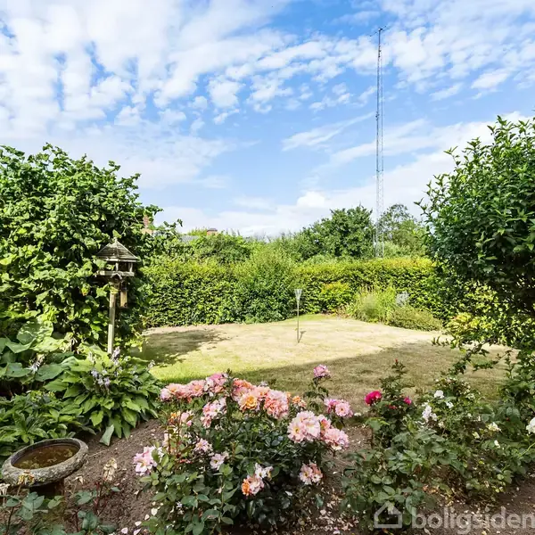 En frodig have med blomsterbede domineret af pink og hvide blomster. En græsplæne ligger i midten, omgivet af grønne buske og træer. Himlen er delvist overskyet.
