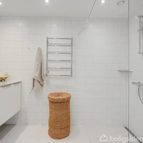 En moderne badeværelseskurv i flet står på gulvet nær et bruseområde med glaspanel. Vask med spejl og håndklædeholder hænger på væggen. Atmosfæren er minimalistisk.