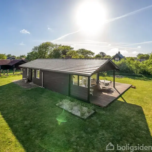 En træhytte står på en velplejet græsplæne under en klar, solrig himmel. Hytten har en terrasse med havemøbler, og omgivelserne er grønne og frodige med spredte træer.